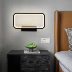 Applique a forma di cornice nera luminosa con staffa rettangolare fissata a una parete grigia. Un comodino grigio scuro con una piccola pianta verde sotto. Un letto marrone sulla destra.