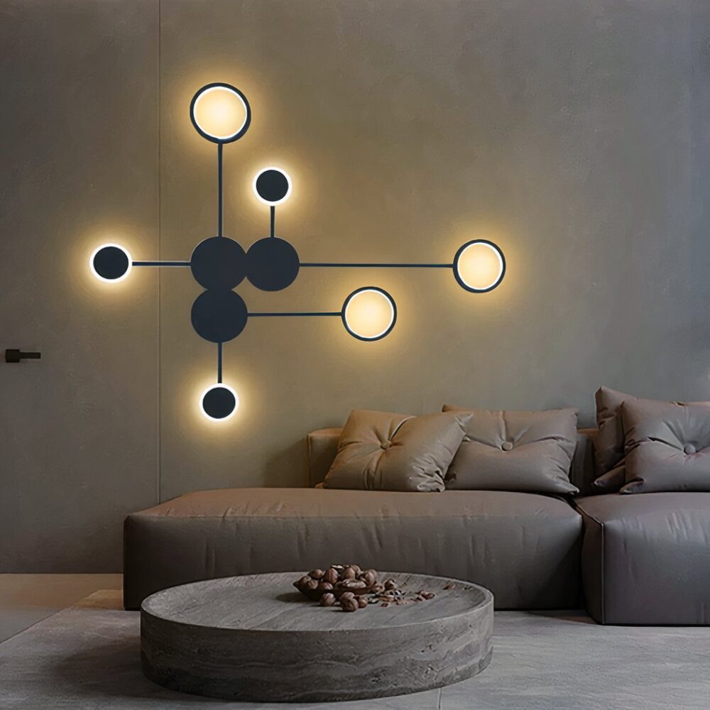 Applique nera composta da cerchi collegati da linee. 3 cerchi neri al centro e 6 cerchi intorno alla luce. Montata su una parete marrone sopra un divano marrone con cuscini coordinati. Tavolino rotondo marrone e tappeto beige sotto.