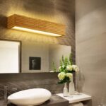 Applique rettangolare in legno accesa e montata su una parete marrone sopra uno specchio rettangolare più lungo della lampada. Sotto lo specchio si trova un lavabo bianco a forma di ciottolo. Alla sua destra si trovano fiori bianchi in sottili vasi bianchi. La parete di destra è beige.