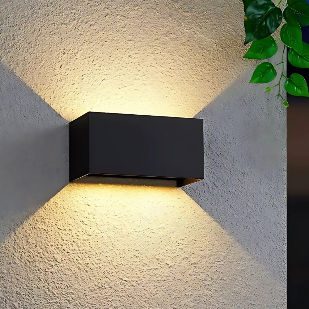 Applique da esterno rettangolare nera a doppio fascio. Montata su una parete esterna beige e accesa.